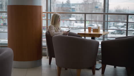 lady seated alone in a cozy mall space, engrossed in her phone, with a busy city view through large windows, the tranquil moment contrasts with the bustling city outside