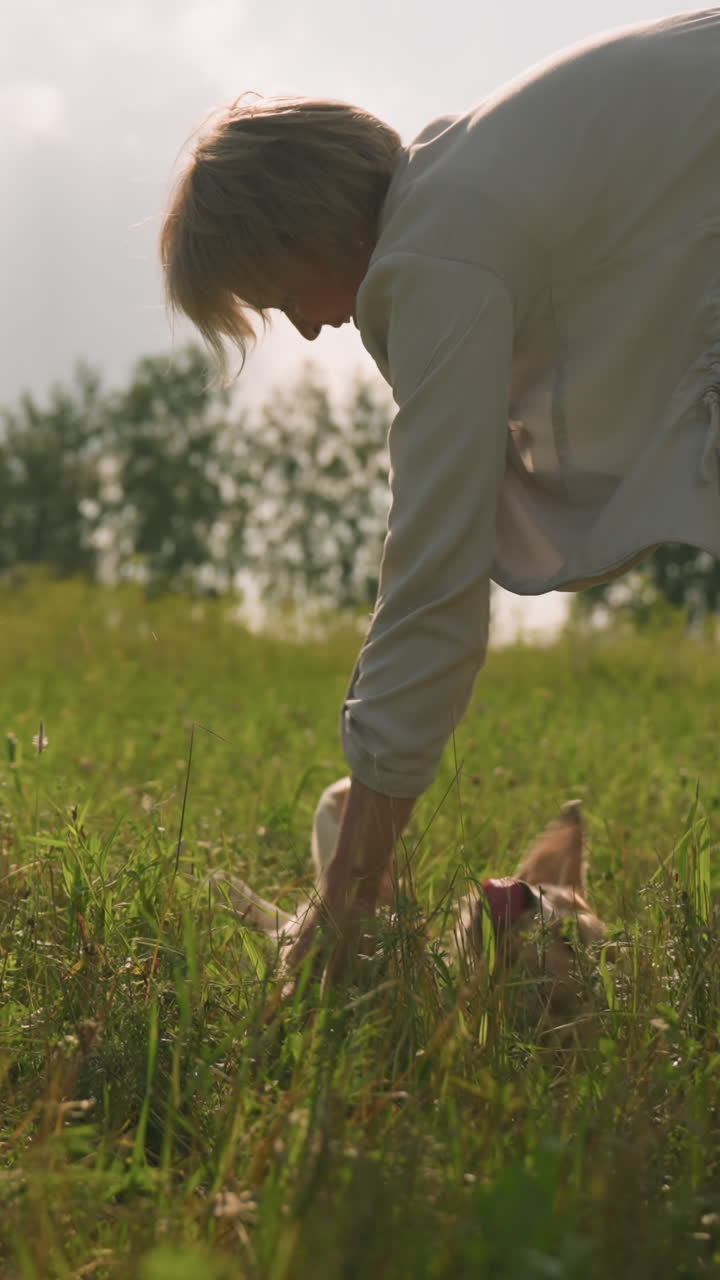 Woman with left hand holding something while playing with her dog as dog happily rolls on grassy field under sunny sky, bright sunlight shines over open landscape