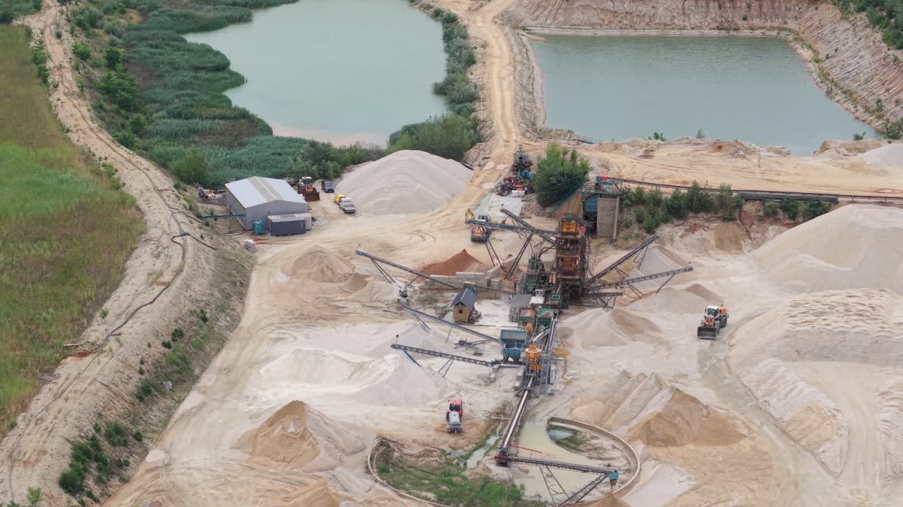 A large sand mining operation with excavators, conveyor belts, and piles of processed materials near artificial ponds. The facility is surrounded by natural vegetation and dirt roads
