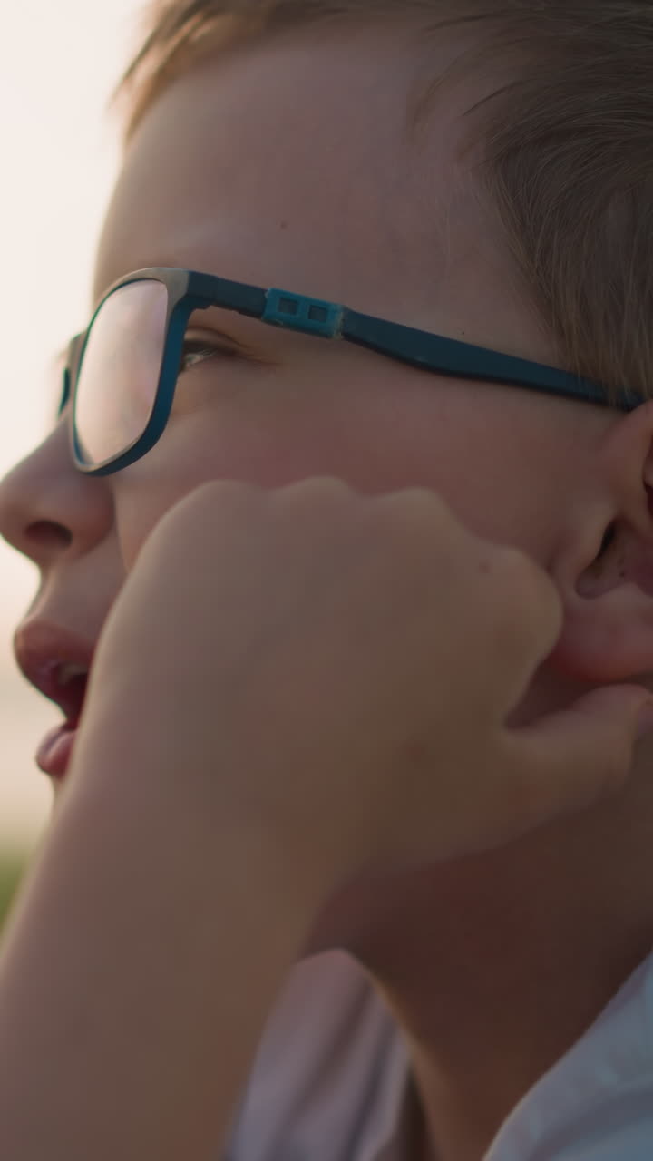 primer plano de un niño con gafas y una camisa blanca, masticando pensativamente con la mano descansada en la mejilla, disfrutando de un momento de paz al aire libre en un campo cubierto de hierba durante la puesta de sol