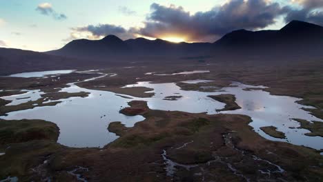 Drohnenaufnahmen-Aus-Der-Luft,-Die-Sich-über-Einer-Moorlandschaft-Aus-Torfmooren-Zwischen-Einem-Mosaik-Aus-Süßwasserseen-Umkehren-Und-Bei-Sonnenuntergang-Auf-Dunkle-Berge-Am-Horizont-Blicken