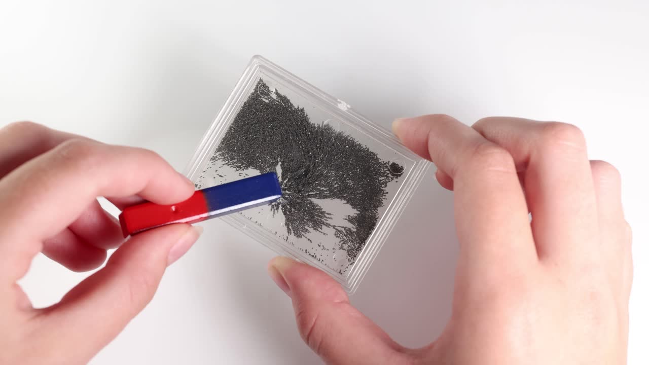 Hands use a magnet to move iron filings in a clear container, demonstrating magnetic fields. Bright lighting highlights the interaction