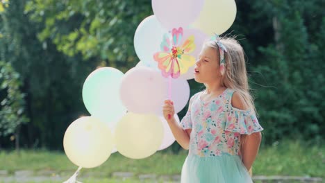 happy little girl in a dress plays with a toy pinwheel. multicolored balloons. children's holiday birthday in nature against the backdrop of the forest