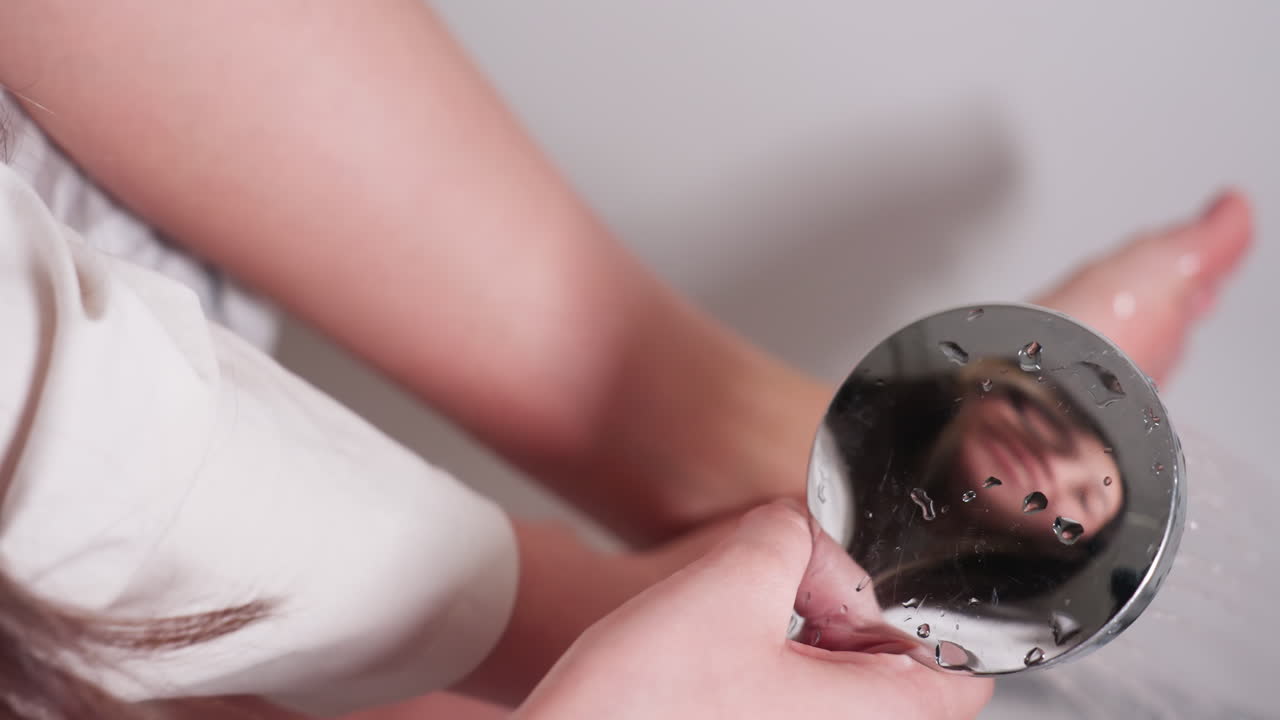 Elegant young girl seated in bath tub washes feet with hand held shower, close view hands guiding stream, face reflection visible on chrome head, calm hygiene routine, soft light over white tub