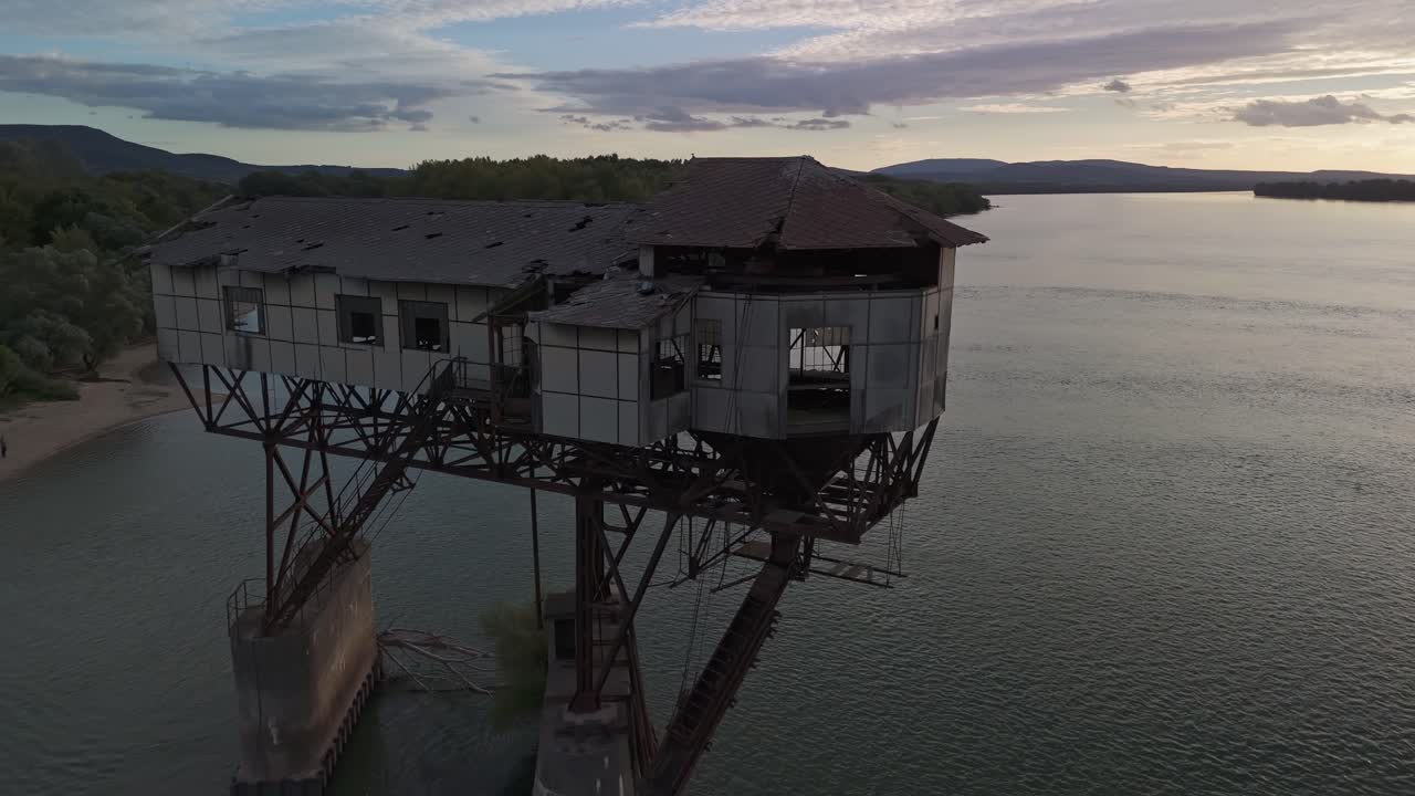 Side tracking close-up drone view of a neglected coal loader over the Danube River in Esztergom, Hungary, showcasing industrial decay and historical heritage amidst a dramatic evening landscape