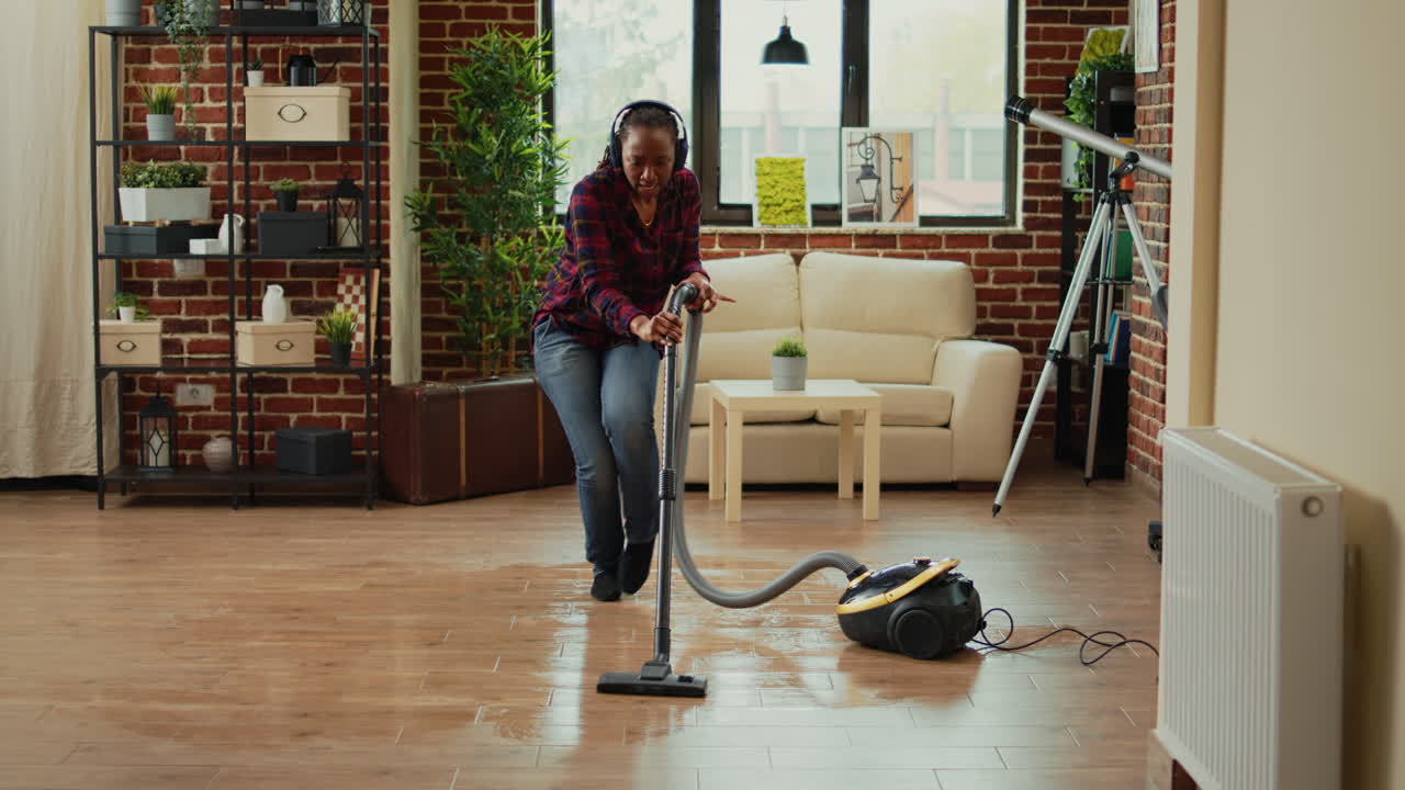 African american housewife using vacuum cleaner and listening to music on headset