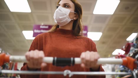 Beautiful-woman-wearing-protective-disposable-medical-mask-and-fashionable-clothes-uses-shopping-cart-while-shopping-in-supermarket