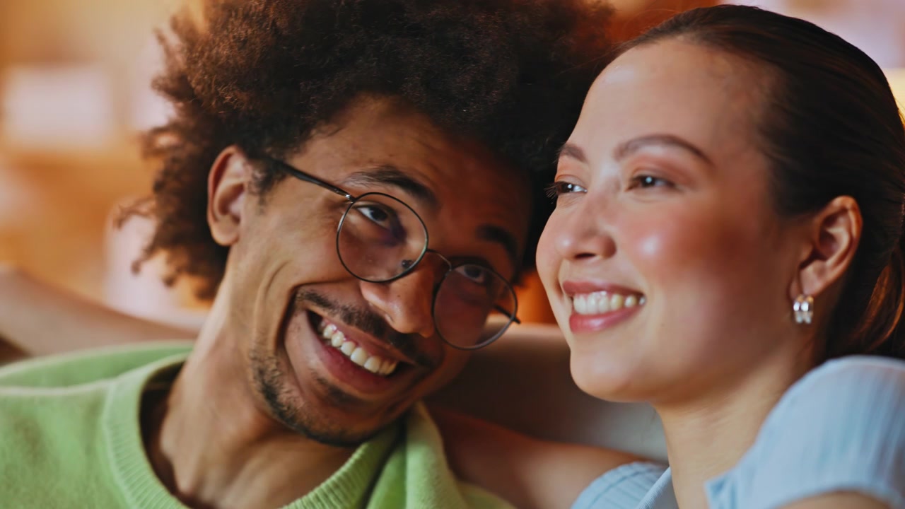 Casal multirracial abraçado, em close, em casa. Homem sussurrando no ouvido da mulher, que ri