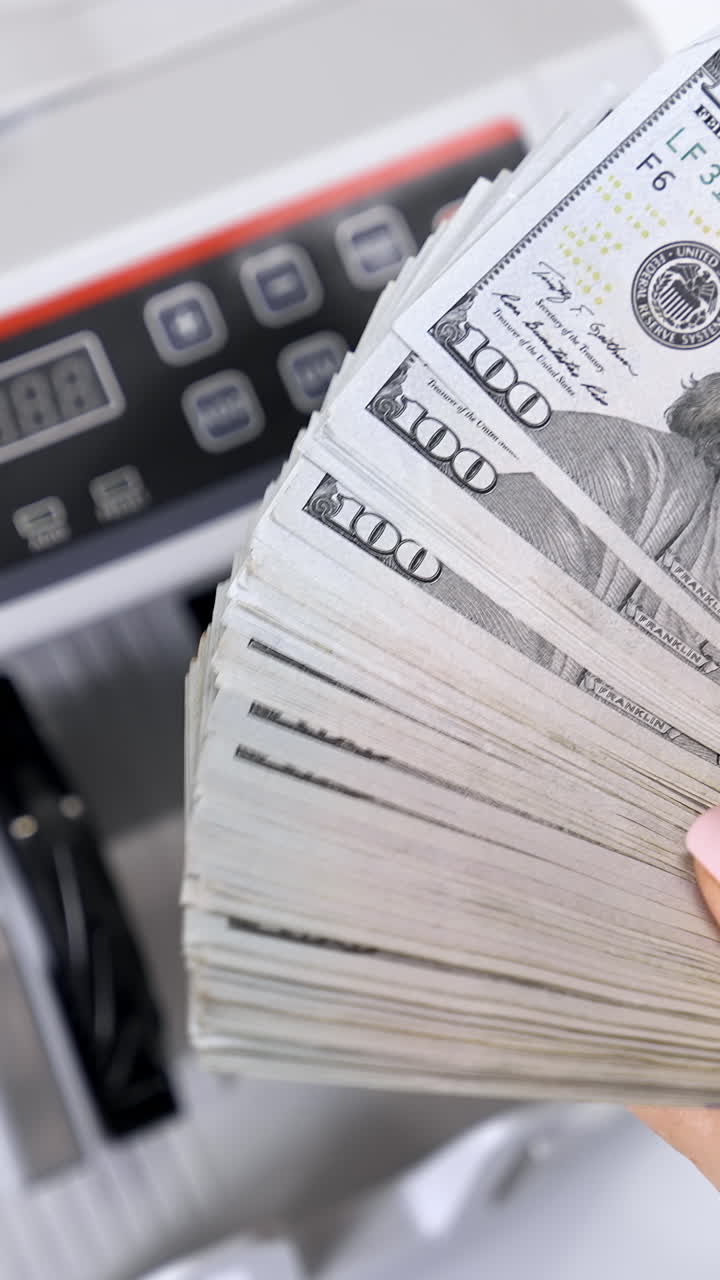 Bundle of one hundred dollar banknotes. Female's hands holding a large sum of money of dollar bills on the background of automatic counting machine. Vertical video