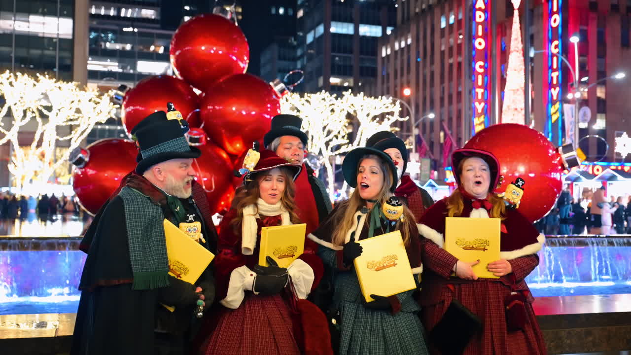 New York, USA, 5 December 2025: Victorian Carolers Singing by Giant Red Ornaments at Night. A group of carolers in traditional Victorian attire performs brightly lit holiday songs near giant red Christmas balls