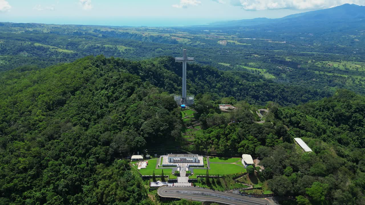 Wide pull‑out aerial revealing the monumental Mt. Samat National Shrine, showcase its towering cross framed by lush forests, valleys, and distant mountains in Pilar, Bataan, Philippines