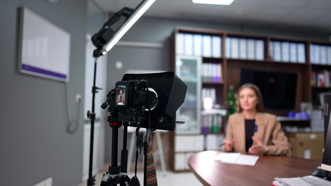 Professional camera with display set on tripod records a video. Woman in beige jacket talks gesturing. Blurred backdrop.