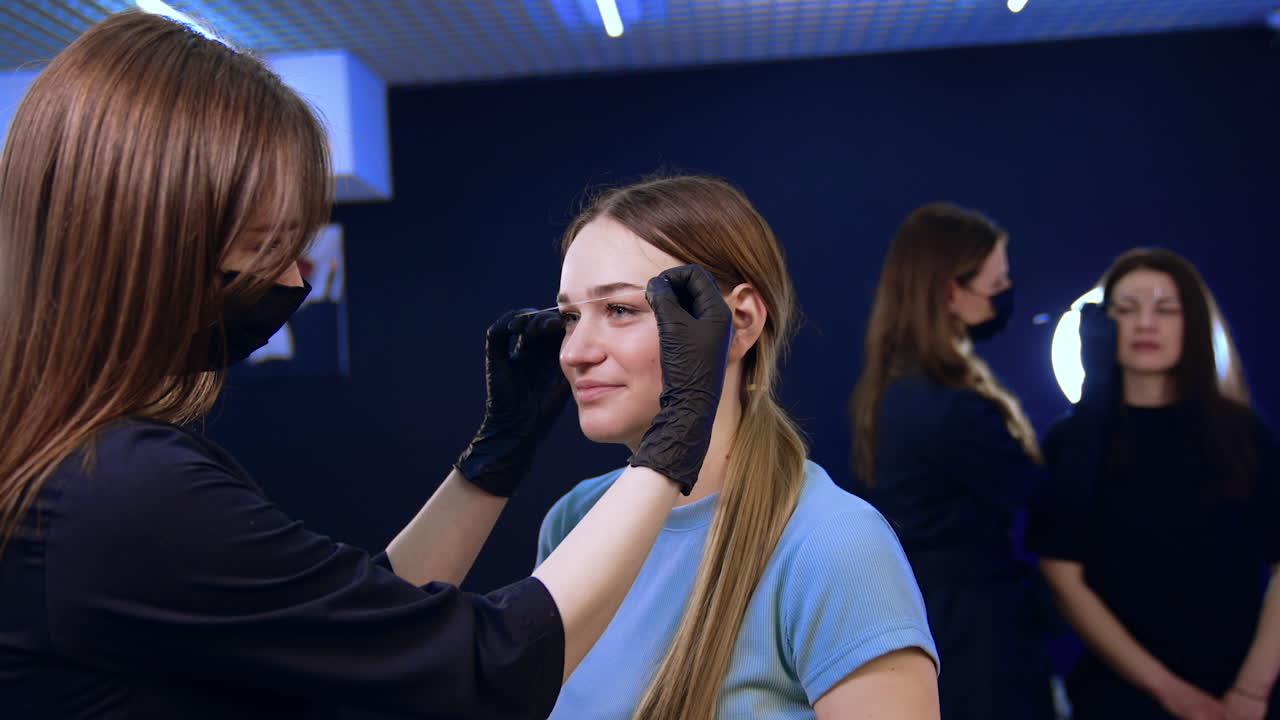 Brunette long-haired cosmetologist draws white lines for eyebrows with the help of a thread. Brow masters work in the salon.