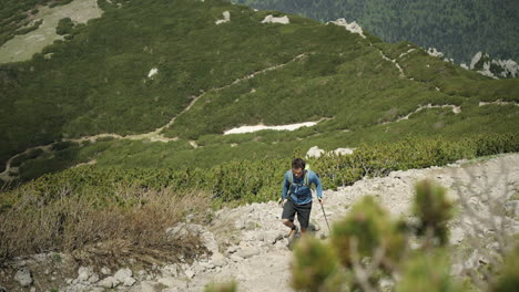 Man-hiking-up-on-a-rocky-path-with-hiking-poles-in-background-is-a-beautiful-green-karst-valley-with-visible-off-road-paths