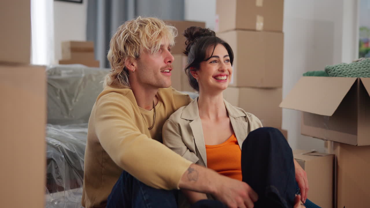 Happy Young Couple Sitting on Floor Among Moving Boxes in New Home
