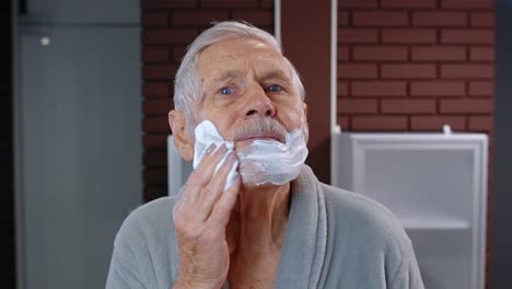 Senior-man-grandfather-in-bathrobe-shaving-with-manual-shaving-blade,-looking-into-mirror.-POV-shot