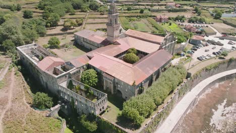 Toma-Aérea-Inclinada-Hacia-Arriba-De-Un-Monasterio-Histórico-Dañado-Ubicado-En-Santa-Maria-De-Oia-Con-La-Vista-De-Algunos-Autos-Estacionados-En-El-Fondo