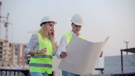 Trabajadores-Con-Dibujos-En-El-Sitio-De-Construcción.-Dos-Trabajadores,-Hombre-Y-Mujer,-Con-Sombreros-Protectores-Trabajando-Con-Dibujos-En-El-Sitio-De-Construcción-Al-Aire-Libre.