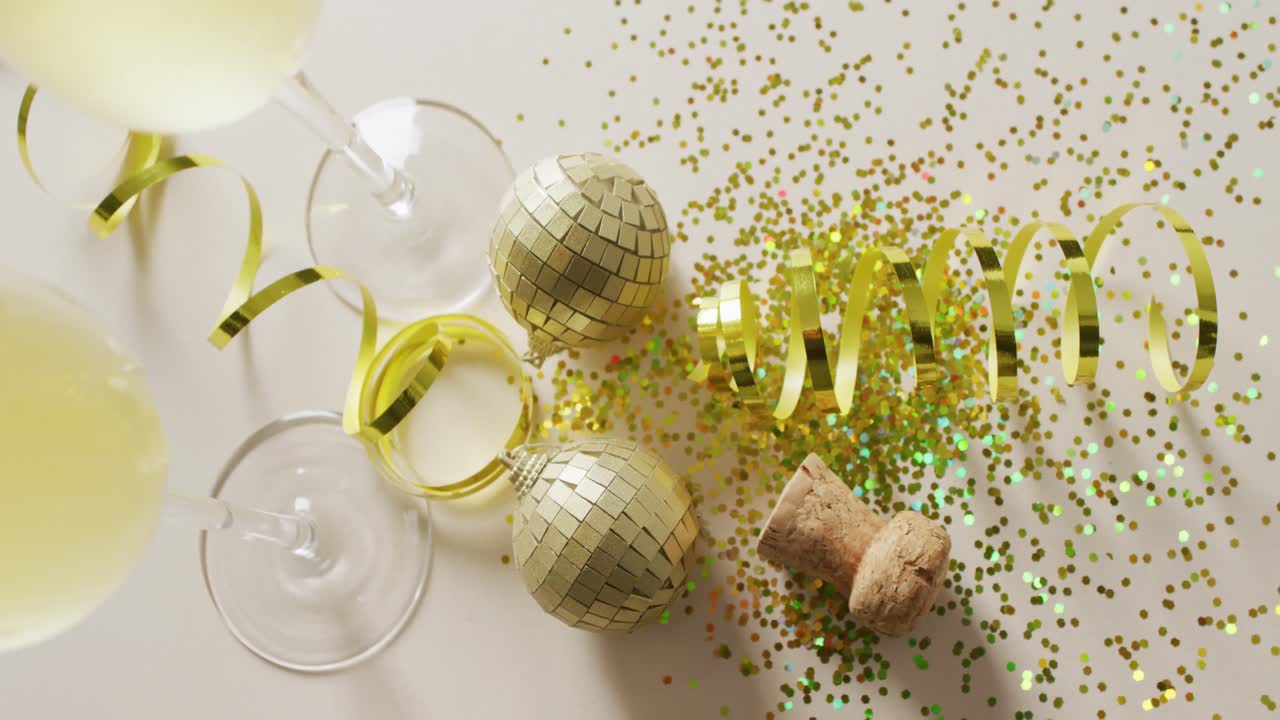 Video dall'alto di due bicchieri da champagne e tappo con stelle filanti dorate, glitter e addobbi su sfondo bianco