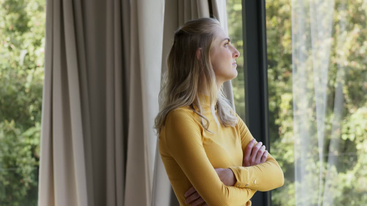 Gazing upward at window, woman in mustard sweater is observing trees, crossing arms, softening gaze