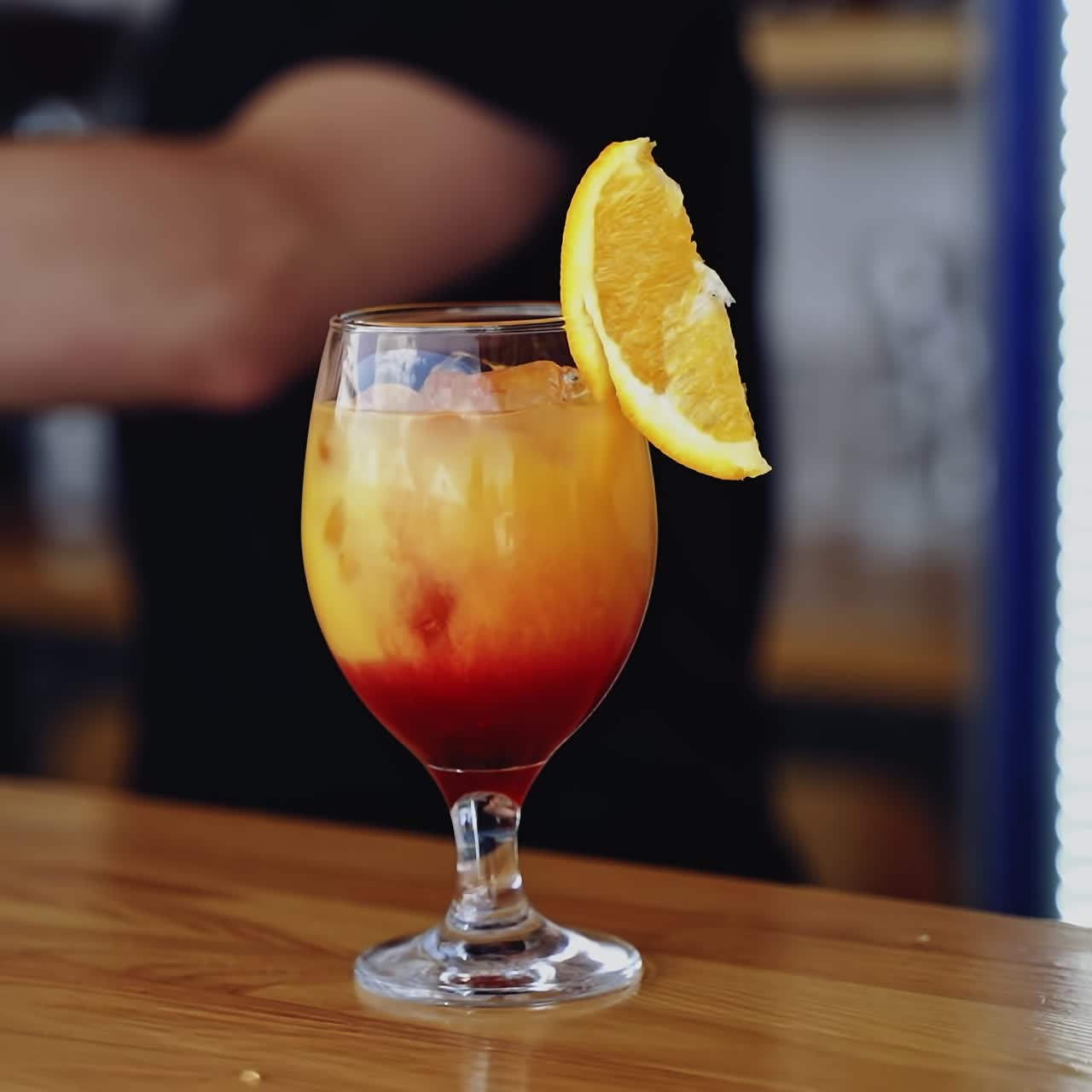 Ready cocktail on a bar table. Bartender puts a slice of orange on the top of the glass with bright beverage. Glass with drink in the nightclub.