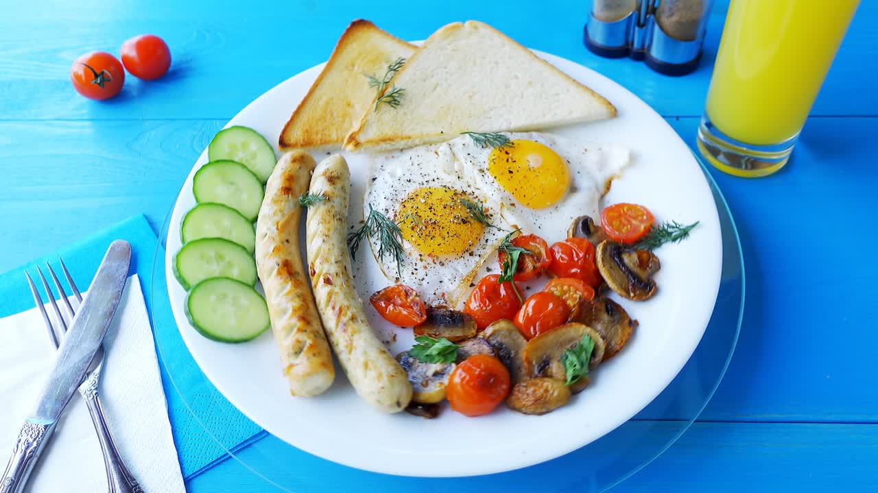 4k loop video of a breakfast of fried eggs, mushrooms, tomatoes, sausages and bread, rotating on a white plate on a wooden blue table.