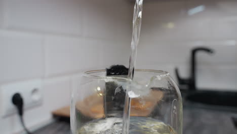 close-up of water being poured into a glass jar in a kitchen setting, the transparent jar captures the motion of the flowing liquid, the blurred background features a sink, countertop, and tiled wall