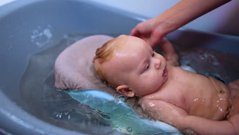 mom's hand is washing hair to her lovely child. beautiful baby boy lies in a blue bath moving hands and feet.