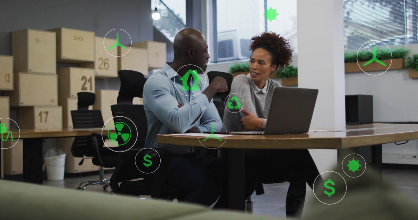 Business duo tapping laptop icon and discussing sustainability plan while energy icons drifting