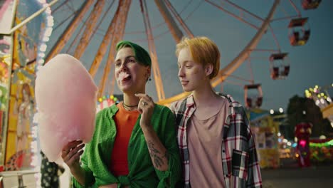 A-girl-with-a-short-green-haircut-in-a-serious-shirt-goes-to-the-half-park-with-her-girlfriend-a-blonde-with-a-short-haircut-in-a-checkered-shirt-and-eats-a-huge-pink-cotton-candy-during-her-date-in-a-bright-and-glowing-amusement-park