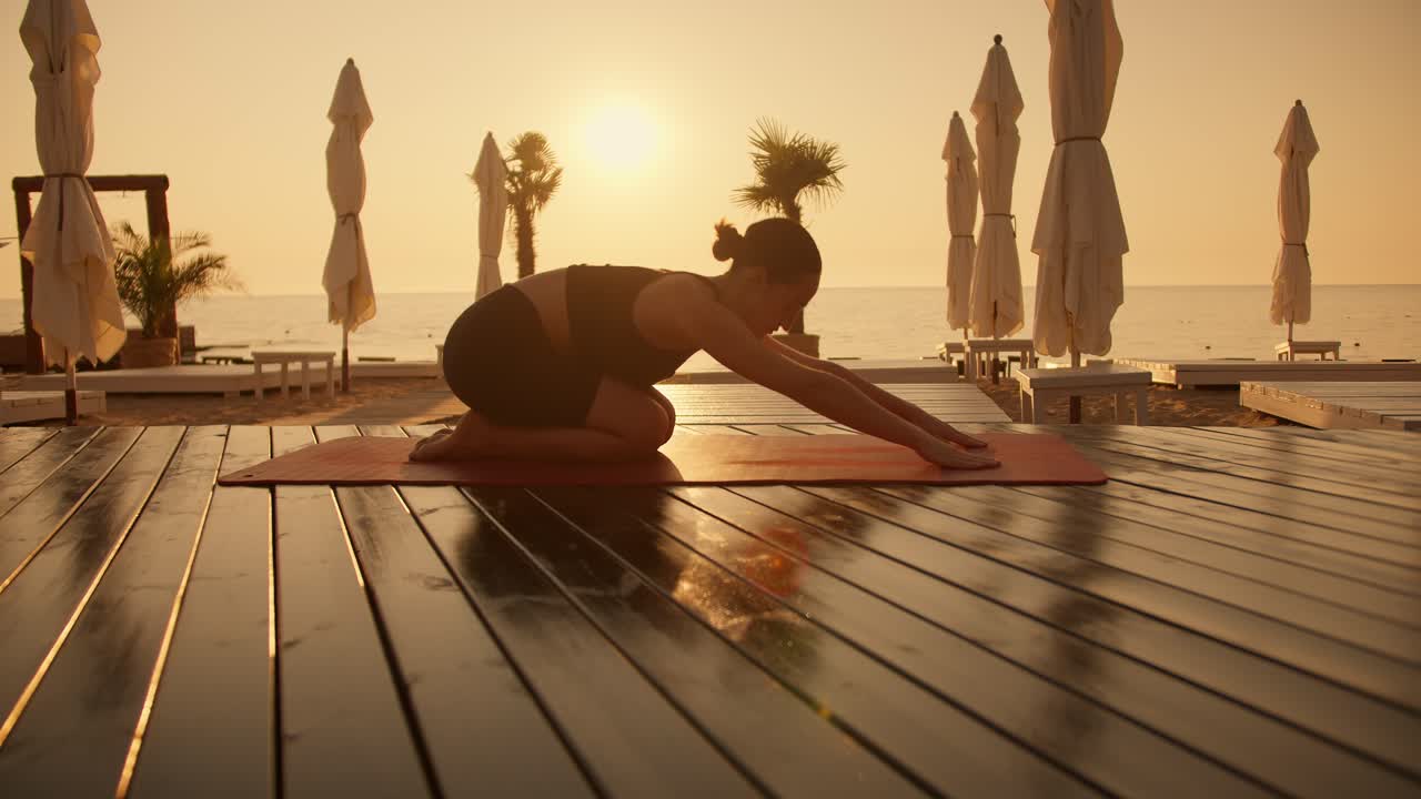 A brunette girl with tied hair in a black sports summer uniform does yoga on red mats on the seashore with a yellow sunrise. The girl takes Half-Tortoise Pose during a yoga class