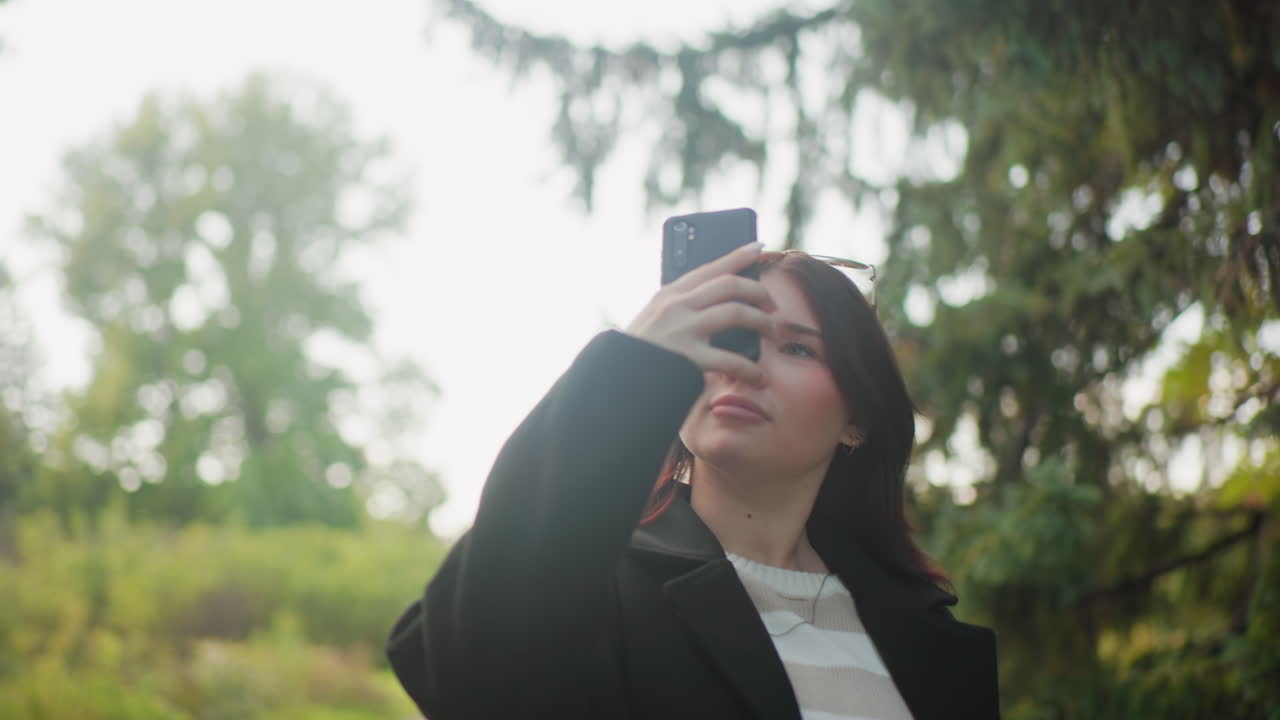 Turista con estilo y abrigo de invierno paseando por un arboreto con un smartphone, deteniéndose bajo árboles de hoja perenne, mirando hacia arriba y encuadrando la escena para hacer fotos, ángulo bajo cinematográfico entre un exuberante follaje y el cielo abierto.