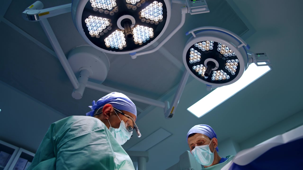 Two medics work in the surgery room under the bright round lamps. Low angle view on the doctors performing operation.