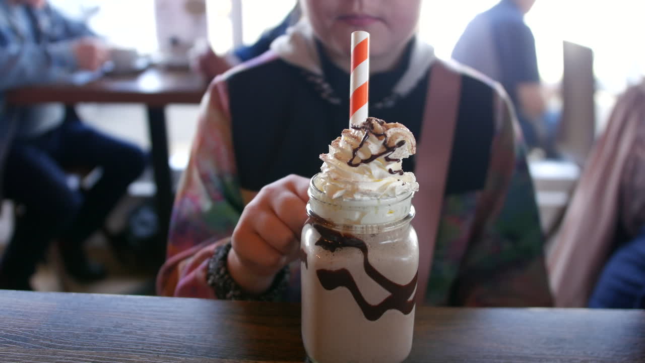 una chica bebiendo de un frasco de albañil lleno de un batido de crema con una copa de batido rociado con jarabe de chocolate
