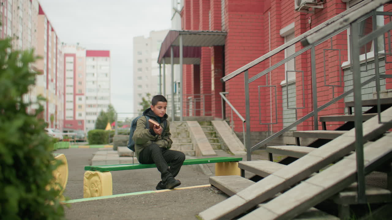 Student carrying backpack and holding book walking toward bench near red brick building, surrounded by green bushes and residential apartments, leaves swaying gently with wind
