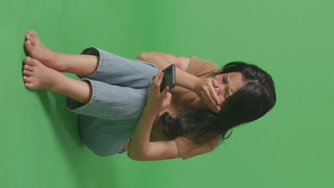 full body of young asian woman victim of violence with bruise on body looking at smartphone and crying while sitting in the green screen background studio