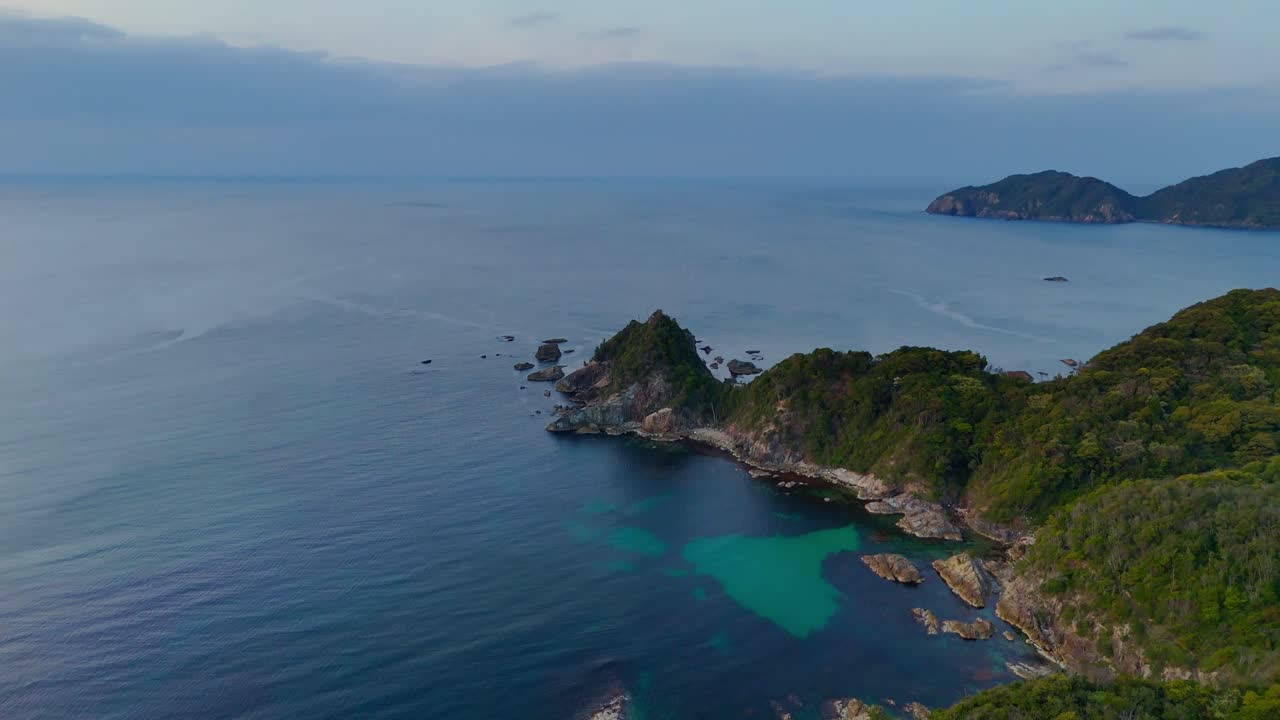 An aerial shot tilts down to reveal a beautiful, rocky coastline. The scene features a hidden cove with vibrant turquoise water, surrounded by lush green hills and the calm, open sea