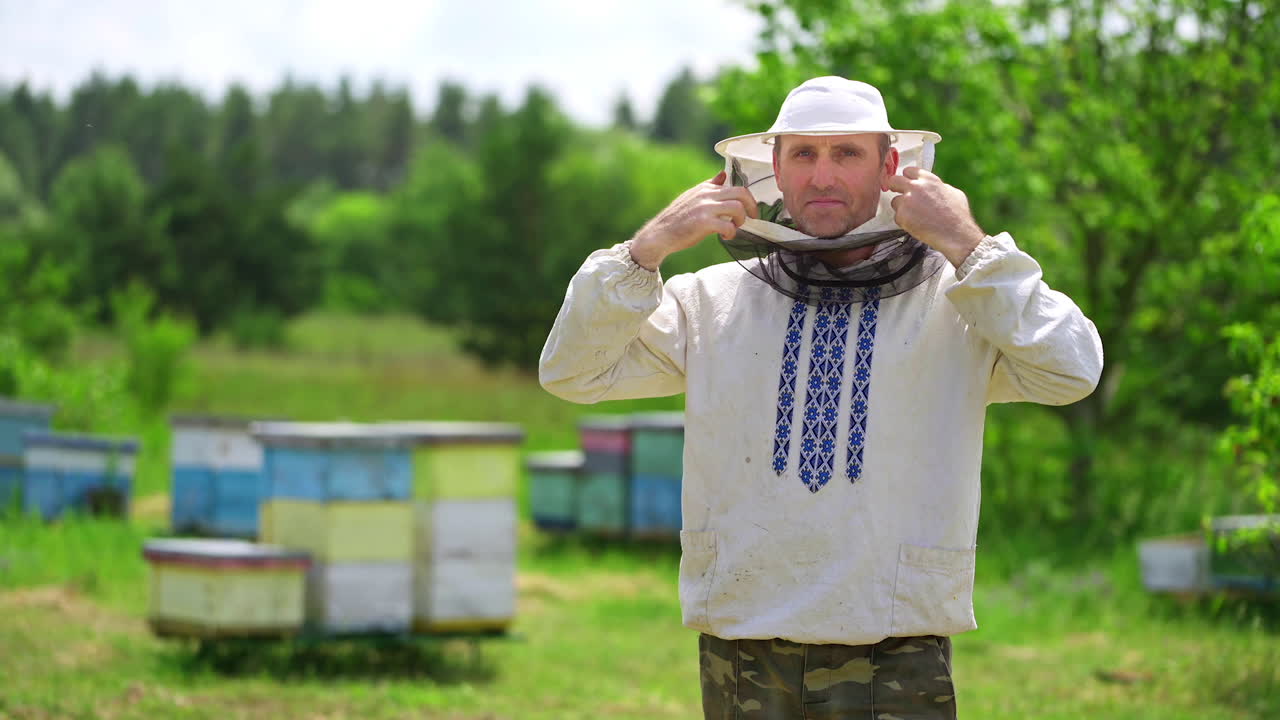 Portrait of beekeeper on apiary. Apiarist wearing protective hat. Man in embroidered shirt puts on hat to protect from bees and smiles on camera.