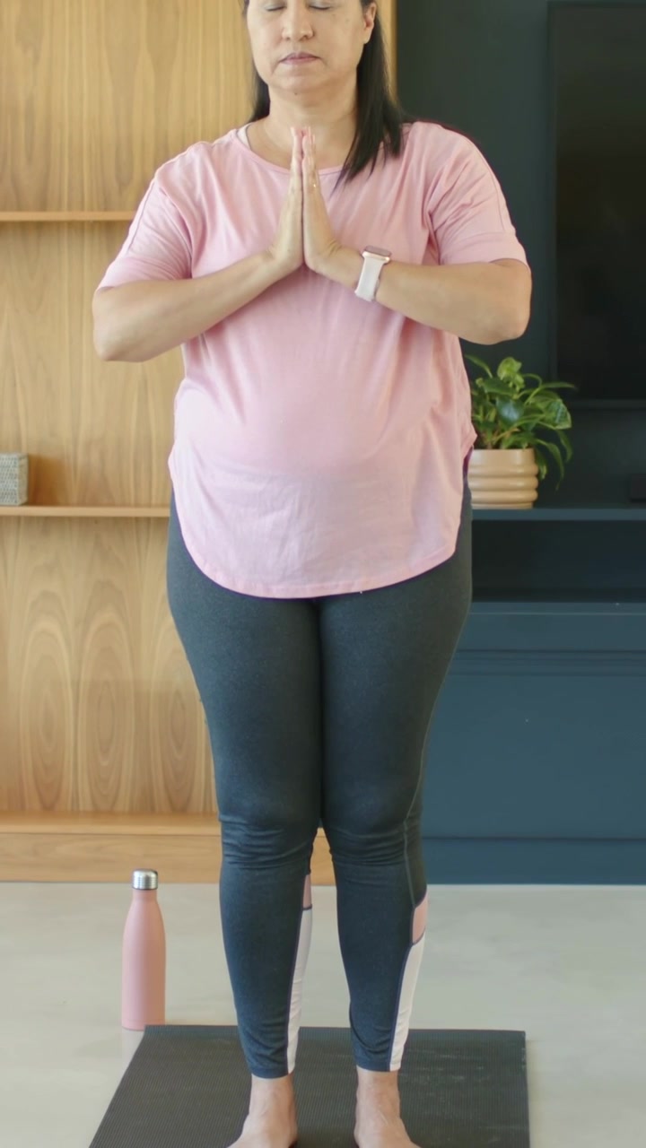 Video verticale: Donna in maglietta rosa con le mani giunte si concentra sul tappetino di casa con una bottiglia.