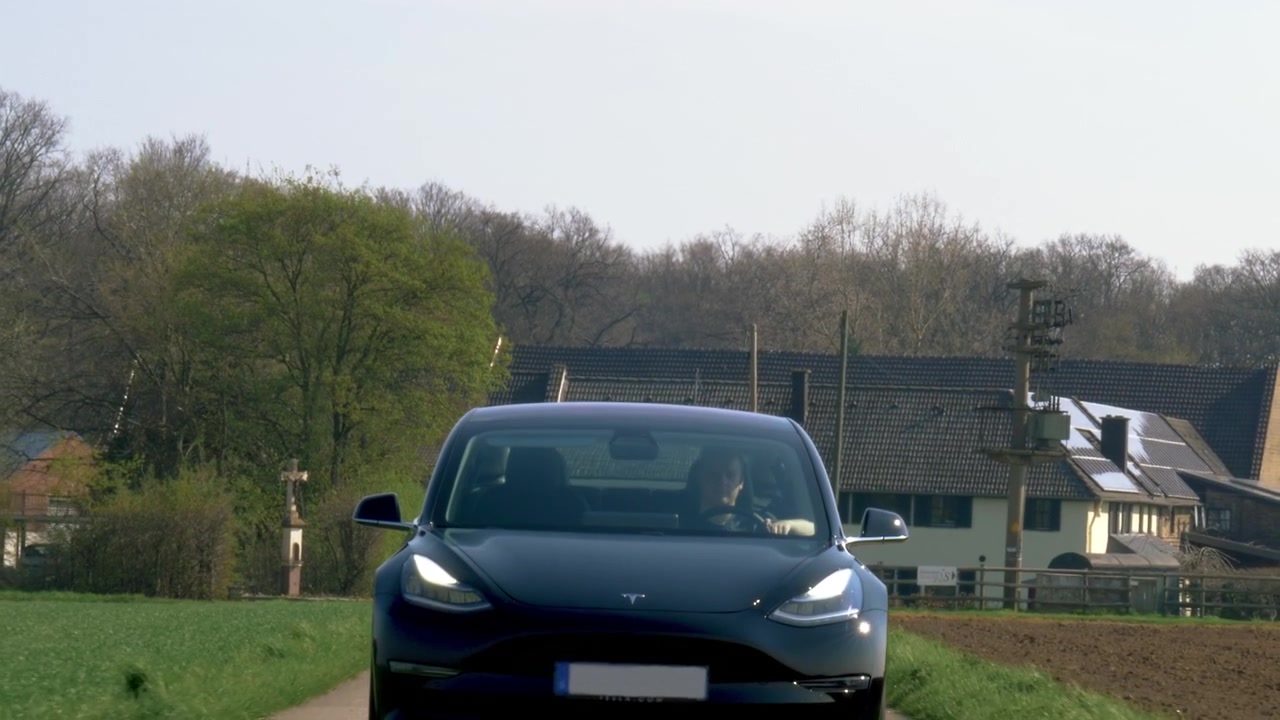 Arka planda küçük bir evle, dar asfalt yolda ilerleyen Tesla Model Üç'ün farklı açılardan çekimleri