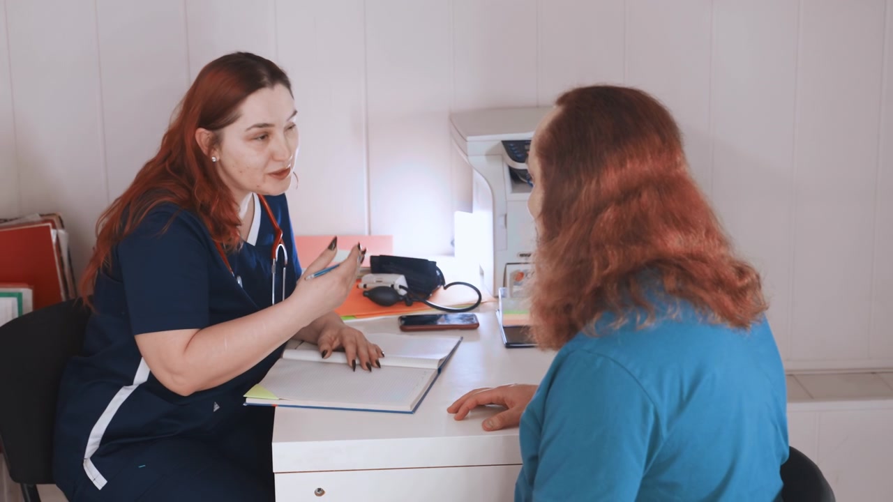 Consulta domiciliar: enfermeira branca à mesa discutindo documentos, estetoscópio visível, contato visual gentil com paciente na sala de estar, luz natural, escuta atenta e avaliação clara para cuidados domiciliares.