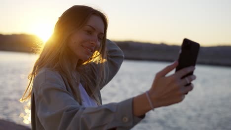 Blonde-woman-looks-at-her-phone-screen-while-standing-on-the-shore,-smiling-happily