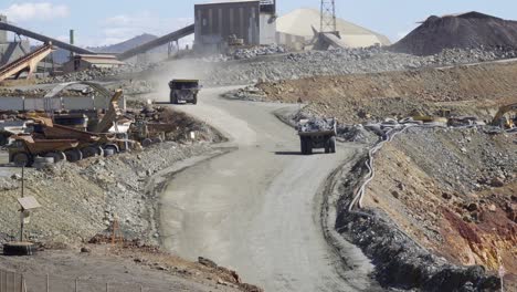 heavy dump trucks transporting ore across the rough tracks of an open-pit mine. The trucks kick up clouds of dust as they load and unload materials