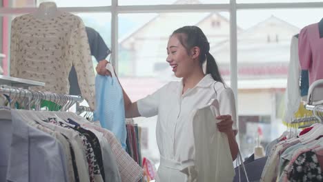 smiling young asian woman with the shopping bags compares two clothing items on hangers while choosing the trendy one at the boutique
