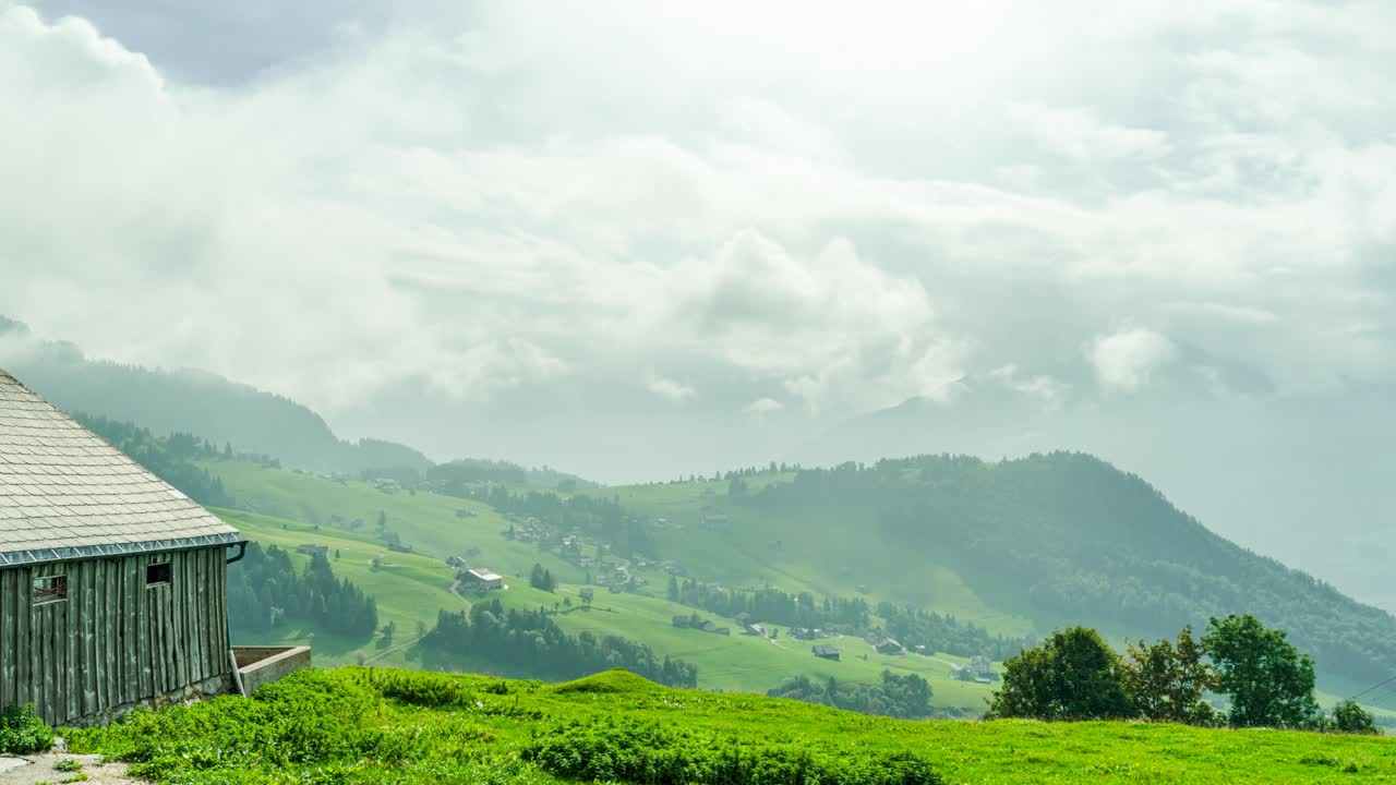 A wooden farmhouse overlooks the rolling green pastures of Amden, St. Gallen, Switzerland, with misty mountains in the background. The timelapse captures shifting clouds and dynamic light changes.