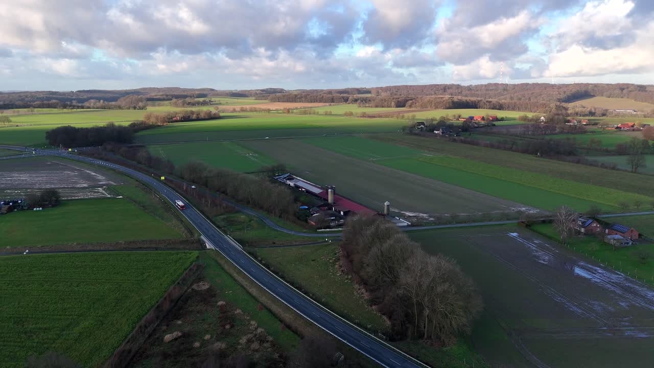 Aerial flight over green cultivated fields and farm house with silo in rural area. Sunset time with sun rays at horizon during cold winter day in USA. American Countryside with bus on intersection.