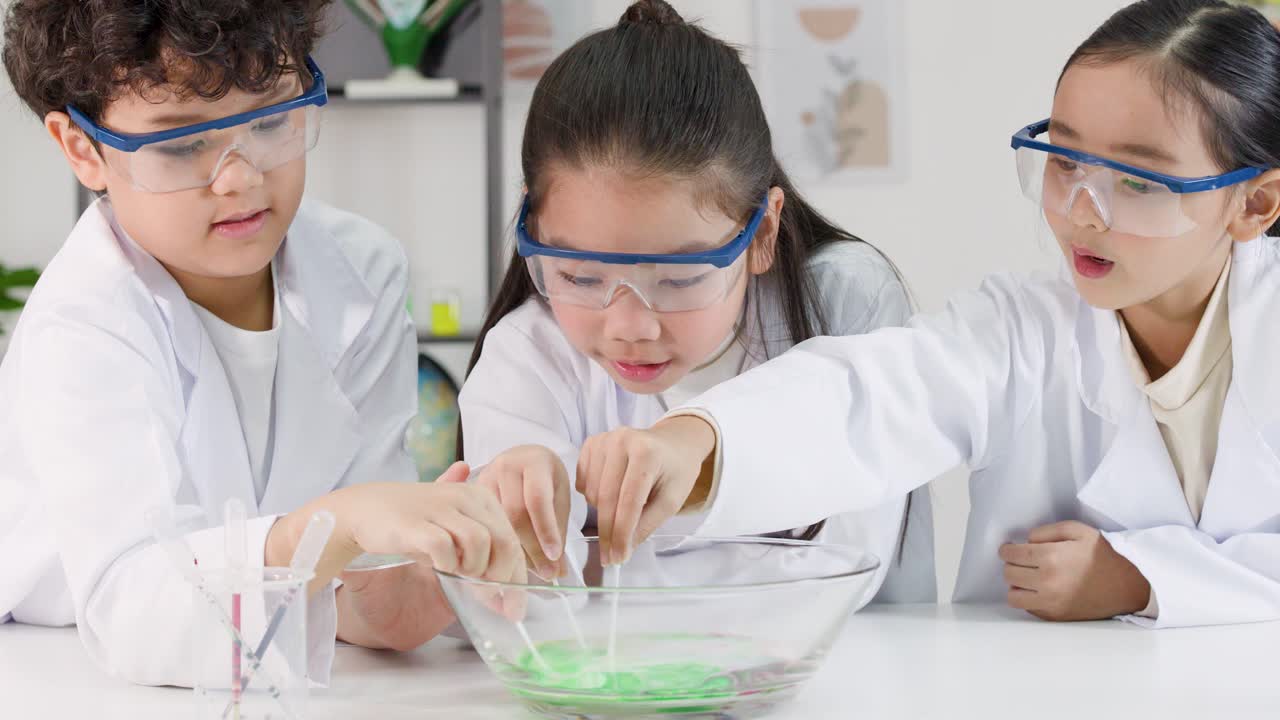 Tiga anak Asia mengenakan jas lab dan kacamata pelindung melakukan percobaan sains susu yang penuh warna bersama-sama