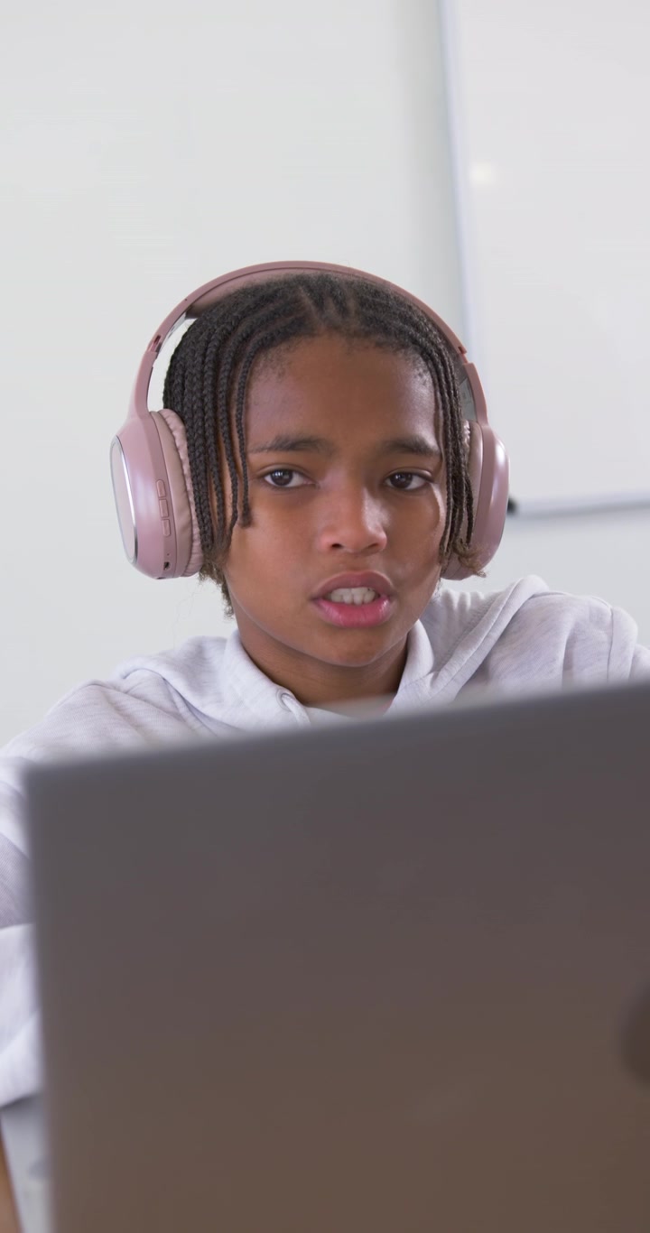 Vertical video: African American teenager with pink headset hearing class leaning at laptop at desk