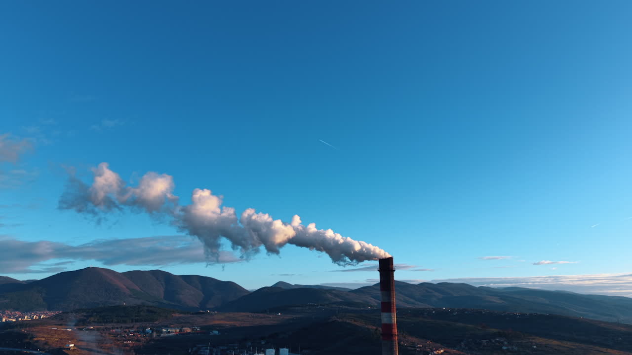 Il fumo si alza dal camino di una fabbrica stagliandosi contro un cielo azzurro terso tra le montagne. Il camino della fabbrica rilascia fumo denso nel cielo sopra le montagne durante le ore diurne.