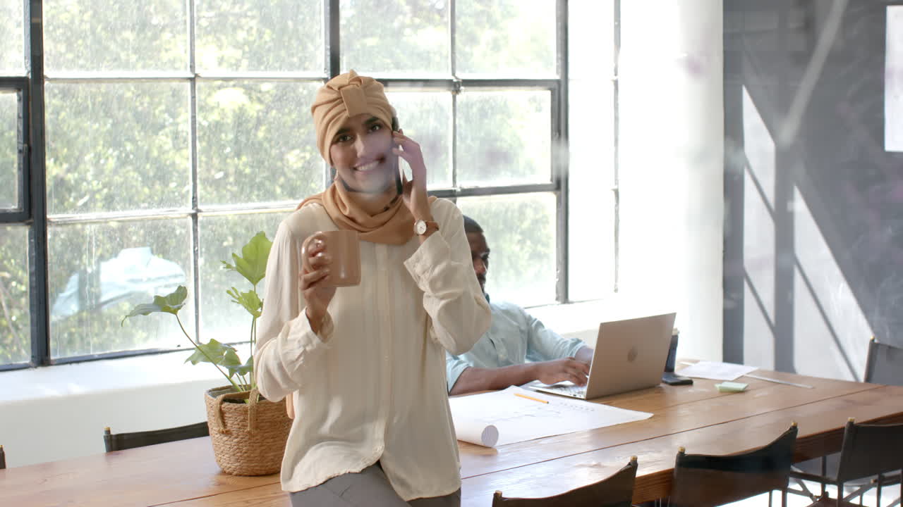 Talking on smartphone and holding coffee cup, woman standing in office workspace
